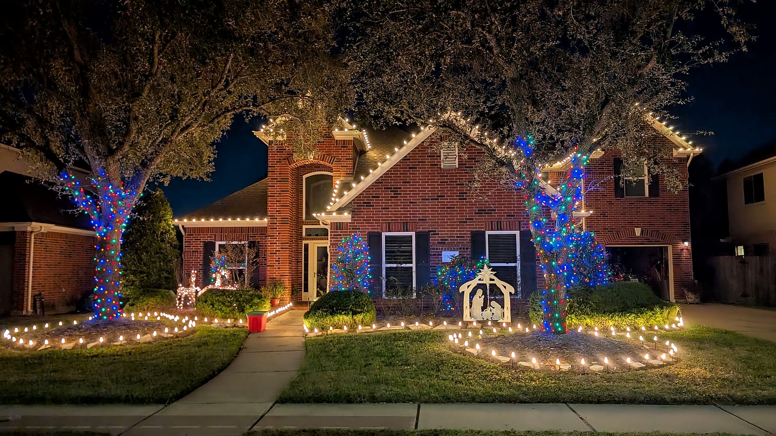 Blue and multicolor wrapped trees with a lit nativity and roofline